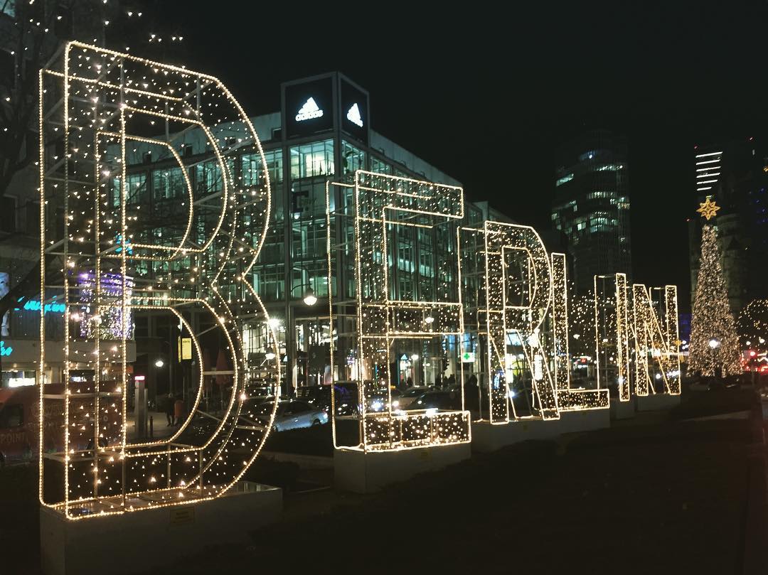 #paracegover Descrição para deficientes visuais: a imagem noturna mostra o canteiro central de uma avenida cdecorado com letras gigantes transparentes, iluminadas por LEDs e formando a palavra Berlin. Ao fundo, uma árvores de natal imensa.