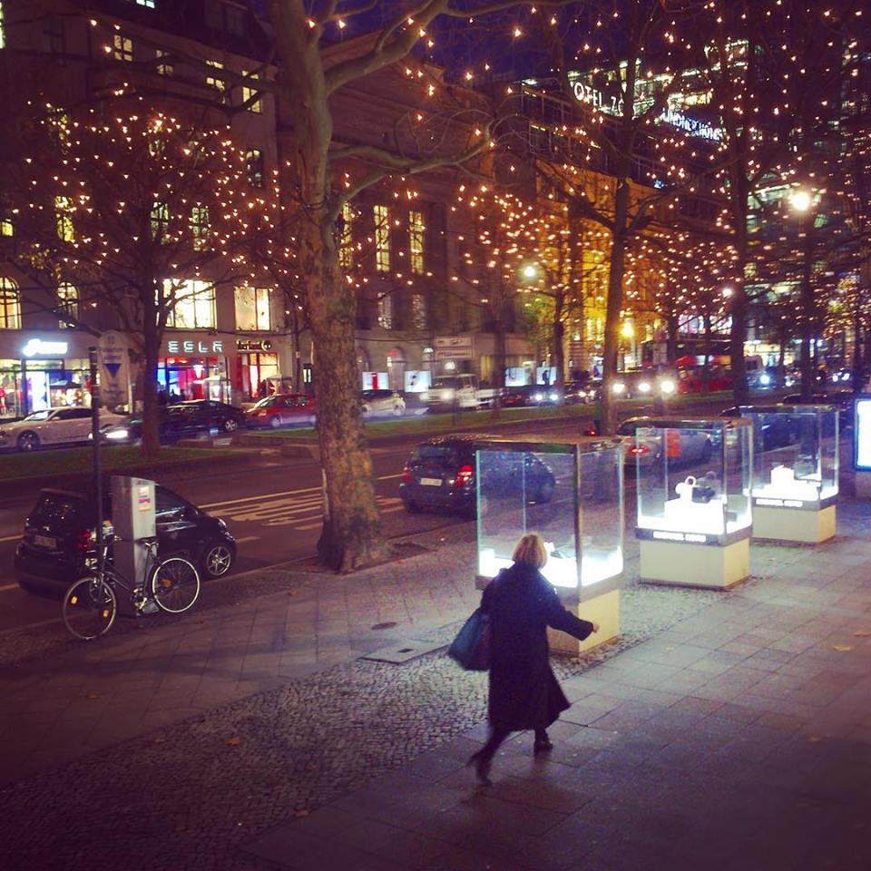 #paracegover Descrição para deficientes visuais: a imagem mostra uma mulher caminhando em uma calçada larga com vitrines de vidro em forma cúbica. Está escuro e as árvores são iluminadas por milhares de pequenas luzes.