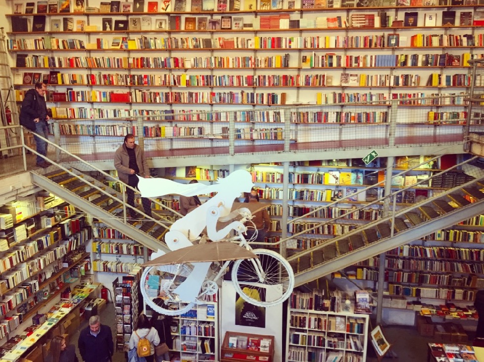 #paracegover Descrição para deficientes visuais: a imagem mostra o interior de uma livraria com suas estantes lotadas em dois pavimentos. Pendurada no teto, uma escultura de uma bicicleta voadora ❤️ — at Livraria Ler Devagar.
