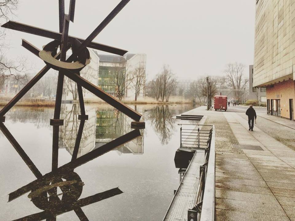 #paracegover Descrição para deficientes visuais: a imagem mostra um homem caminhando em uma calçada, do lado direito. Do lado esquerdo, um lago que reflete a escultura formada por tubos. A névoa toma conta de tudo, forjando um clima de mistério e suspense... — at Theater am Potsdamer Platz.