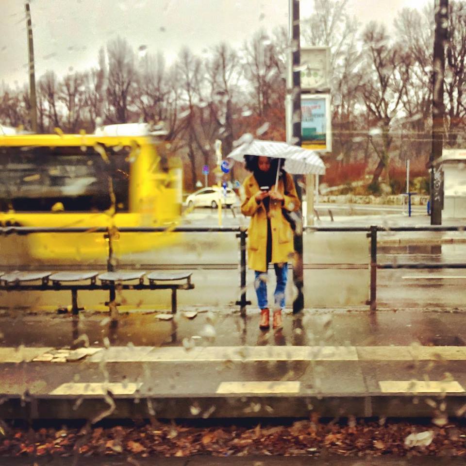 #paracegover Descrição para deficientes visuais: a imagem mostra uma moça de sombrinha branca e capa de chuva amarela esperando o tram e consultando seu smartphone. Acabou de passar um ônibus atrás dela (também amarelo). A cena se passa na frente de um parque em que todas as árvores estão apenas nos galhos. — at Platz der Vereinten Nationen.