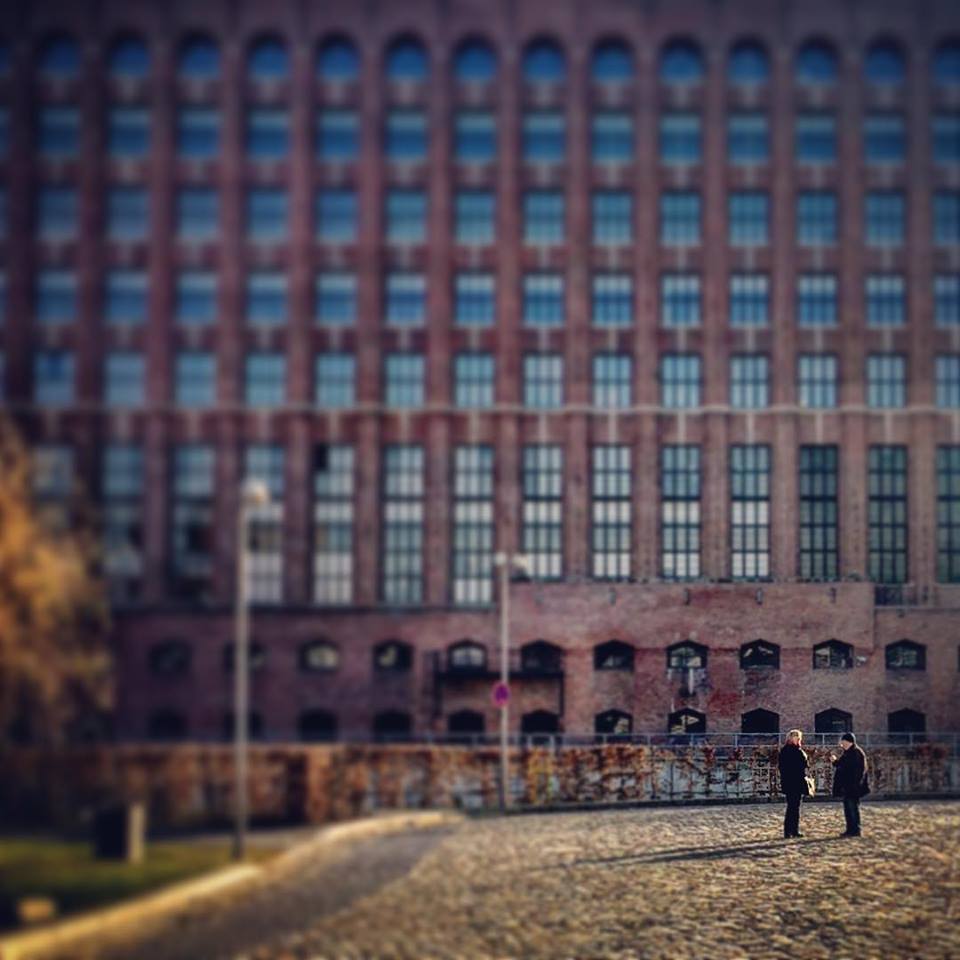 #paracegover Descrição para deficientes visuais: a imagem mostra duas pessoas à distância (em foco) conversando. Atrás delas (desfocado), um maravilhoso prédio industrial cheio de janelas. — at Tempelhofer Hafen.