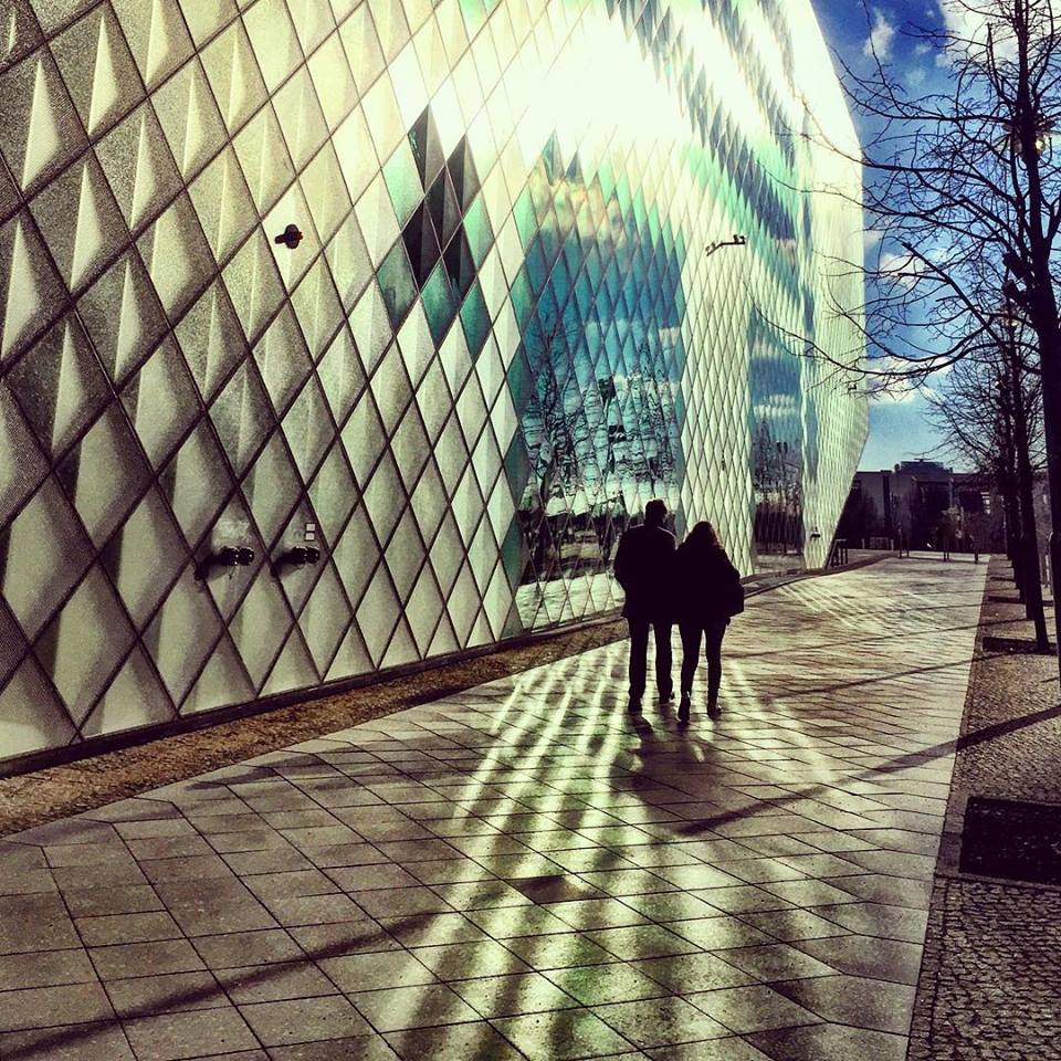 #paracegover Descrição para deficientes visuais: a imagem mostra as silhuetas de um casal caminhando sobre uma calçada. A parede lateral é do centro de exposições Futurium; toda espelhada e com formas geométricas, faz a reflexão do sol parecer uma performance. — at Futurium.