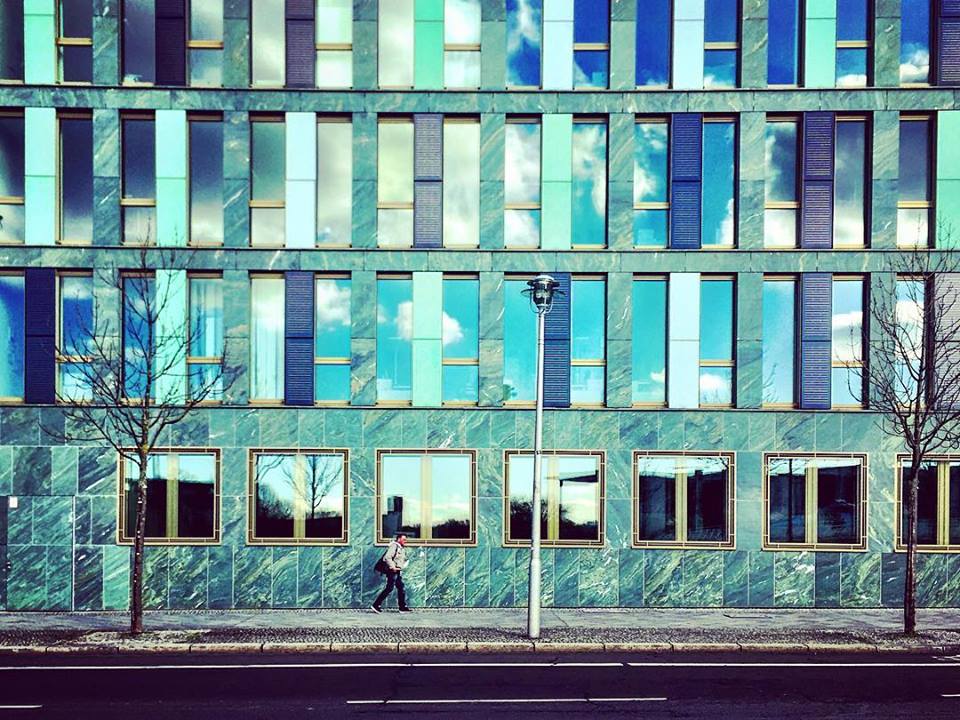 #paracegover Descrição para deficientes visuais: a imagem mostra a fachada de um prédio toda construída em ardósia; as janelas são compridas e refletem o céu. Um homem passa apressado pela calçada. — at Kapelle-Ufer.