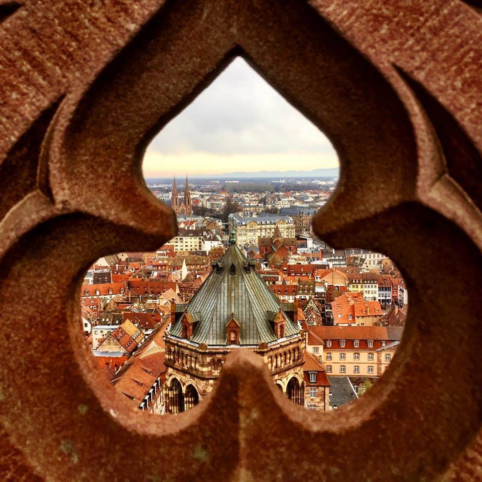 #paracegover Descrição para deficientes visuais: a imagem mostra uma parte da cidade de Strasburgo com seus telhados maravilhosamente inclinados através da fenda em forma de trevo da cerca vazada do pátio de observação.