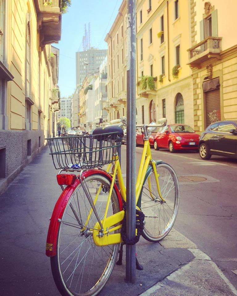 #paracegover Descrição para deficientes visuais: a imagem mostra uma bicicleta com o quadro amarelo e o paralamas traseiro vermelho amarrada a um poste. Há dois carros vermelhos estacionados na rua para combinar. Os prédios são todos amarelos também.