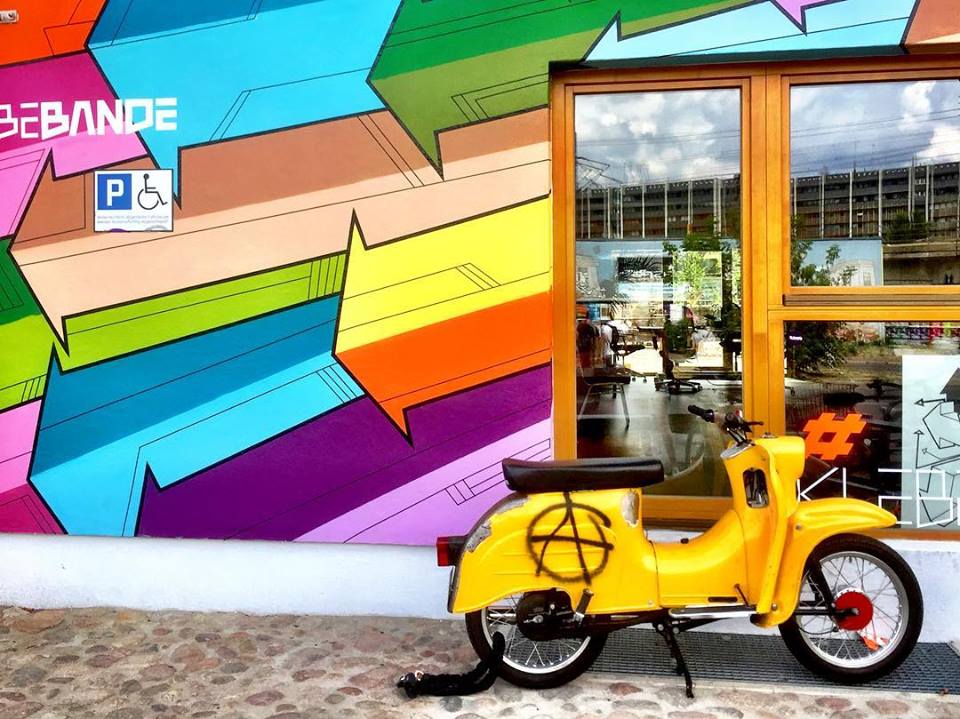 #paracegover Descrição para deficientes visuais: a imagem mostra uma vespa amarela estacionada em frente a uma fachada toda pintada com setas coloridas. — at Holzmarkt25.