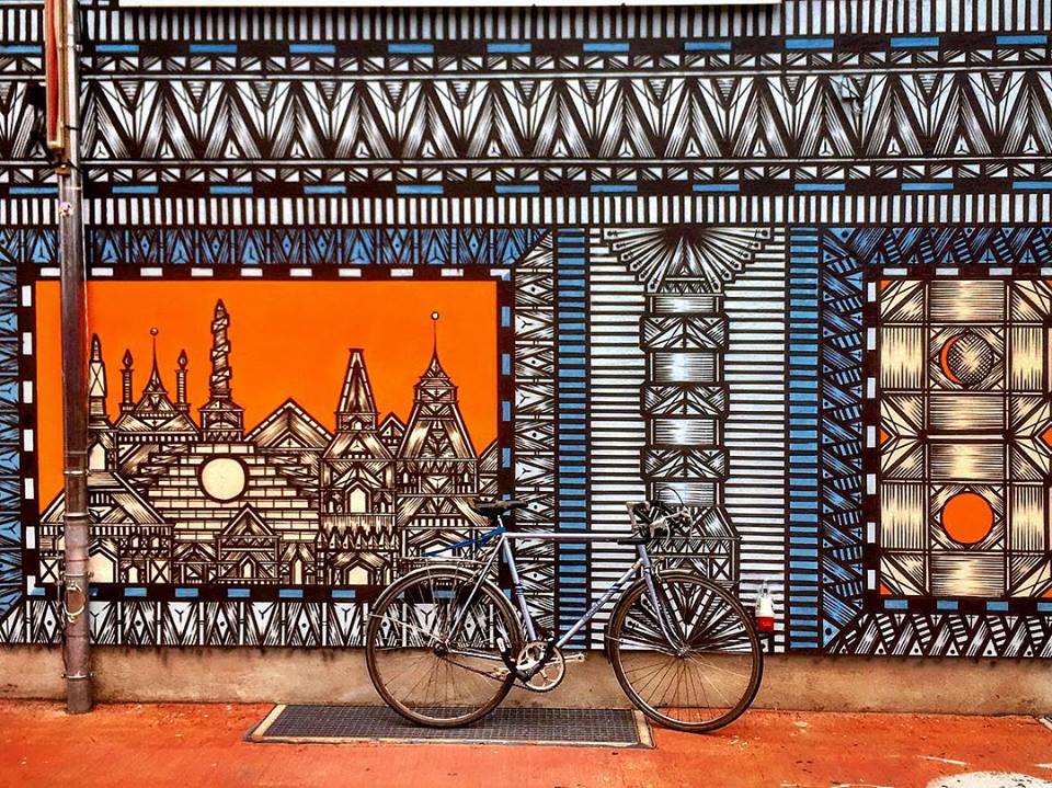 #paracegover Descrição para deficientes visuais: a imagem mostra uma bicicleta estacionada em frente a uma fachada estampada com desenhos incrivelmente detalhados em formas geométricas nas cores azul, laranja e branco. — at Holzmarkt25.