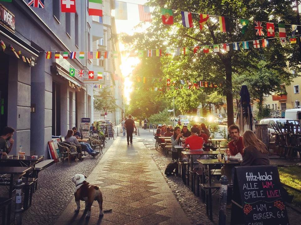 #paracegover Descrição para deficientes visuais: a imagem mostra um bar com mesas na calçada arborizada todo decorado com bandeirinhas de países participantes da Copa. Uma pessoa vem caminhando contra a luz do sol. Em primeiro plano, um pug charmoso. — at Vineria Carvalho Kreuzberg.