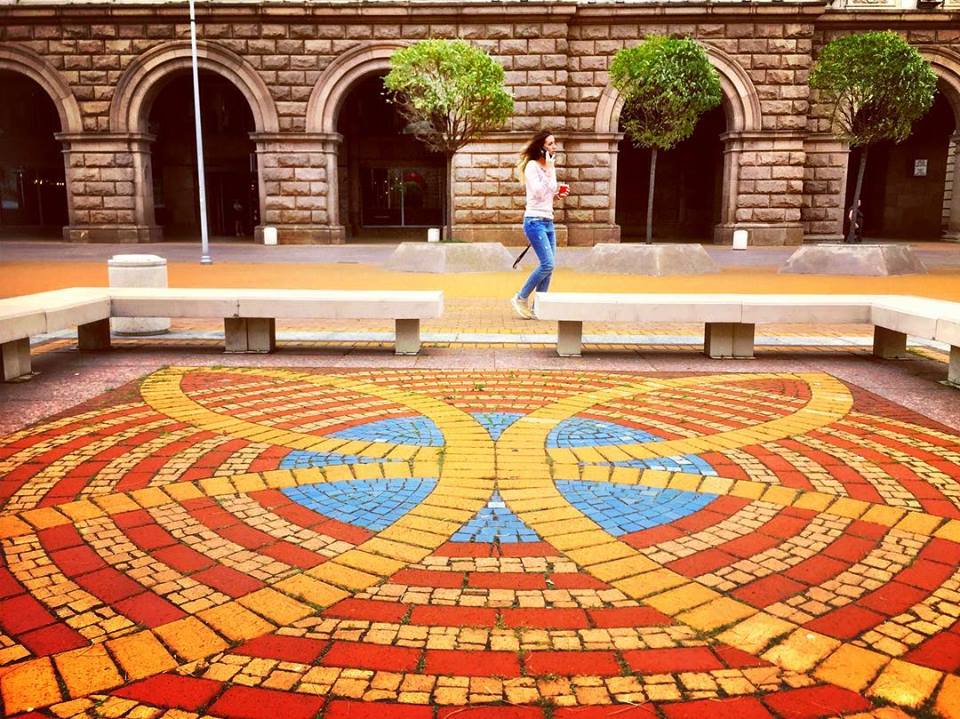 #paracegover Descrição para deficientes visuais: a imagem mostra uma moça caminhando enquanto fala no celular. Em primeiro plano uma calçada com um mosaico nas cores laranja, amarelo e azul. — at Largo di Serdica.