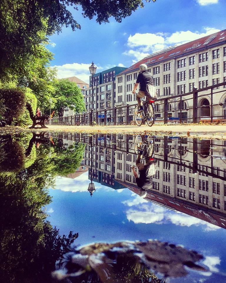 #paracegover Descrição para deficientes visuais: a imagem mostra um ciclista passando pela margem de um dos canais do rio Spree. Ao fundo, prédios e céu azul. A cena é refletida numa poça d’água. — at Spittelmarkt.