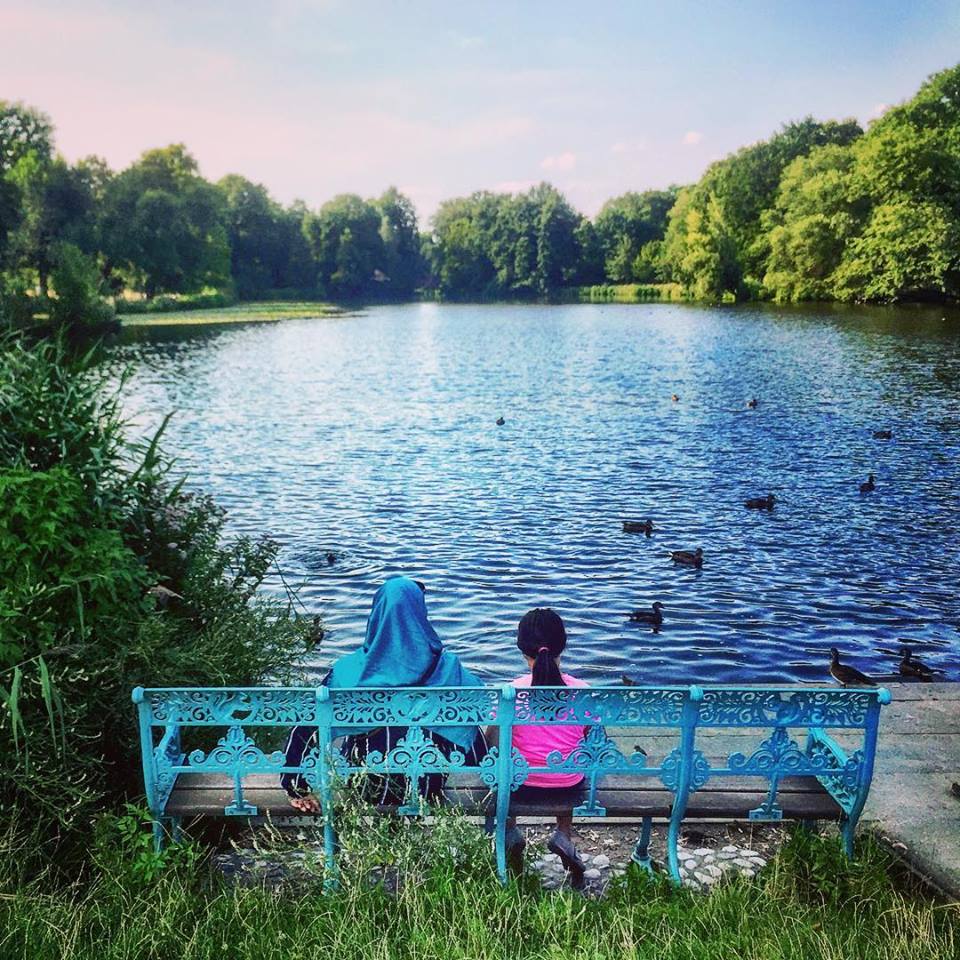 #paracegover Descrição para deficientes visuais: a imagem mostra uma mulher e uma menina de costas, sentadas em um banco de ferro azul. Elas estão contemplando o lago (também azul) dos jardins do Castelo de Charlottenburg. A mulher tem a cabeça coberta com um véu azul. A menina veste uma blusa pink. — at Charlottenburg Palace, Charlottenburg West Berlin.