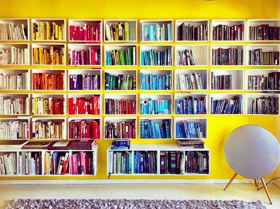 #paracegover Descrição para deficientes visuais: a imagem mostra uma estante de livros formada por 38 caixas de madeira pintadas de branco sobre uma parede amarela (mais 4 caixas para livros maiores da Ikea na parte de baixo). À direita, nicho para a caixa de som em forma de disco com pés de palito. Os livros estão organizados por cores. — in Berlin, Germany.