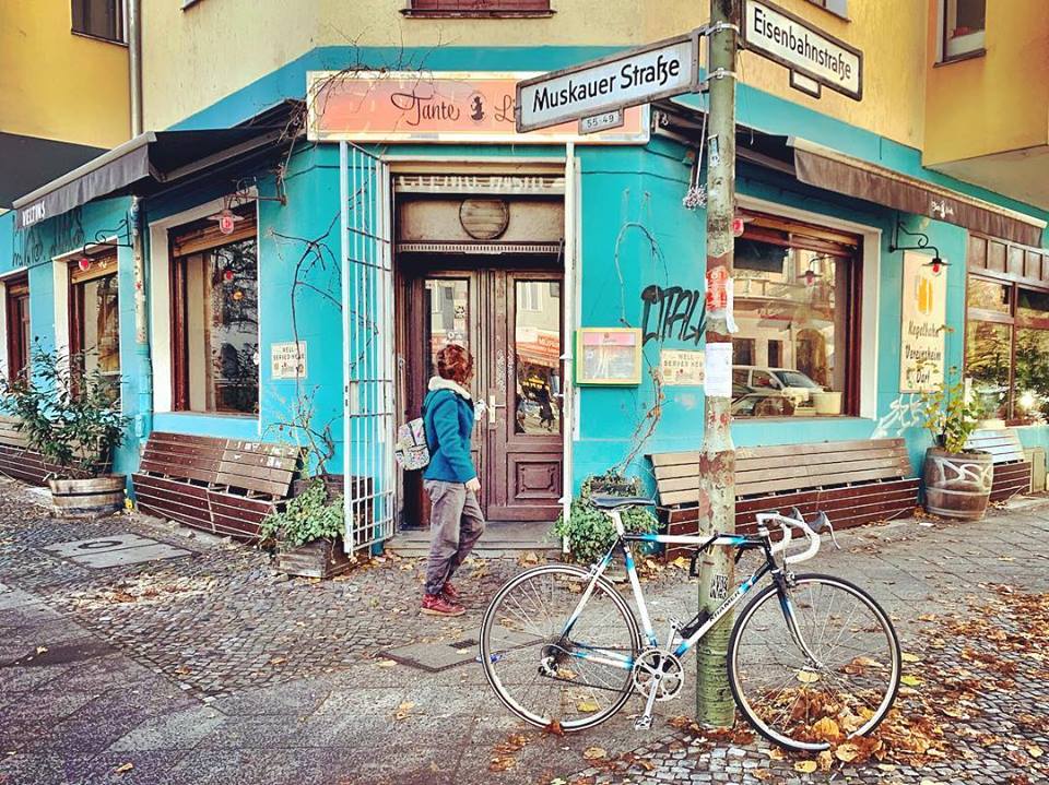 #paracegover Descrição para deficientes visuais: a imagem mostra um café numa esquina da cidade. Há bancos de praça de madeira nas laterais e as paredes são azul turquesa. Em primeiro plano, uma bicicleta estacionada no poste com o nome das ruas. Uma moça está passando pela porta no momento. — at Tante Lisbeth.