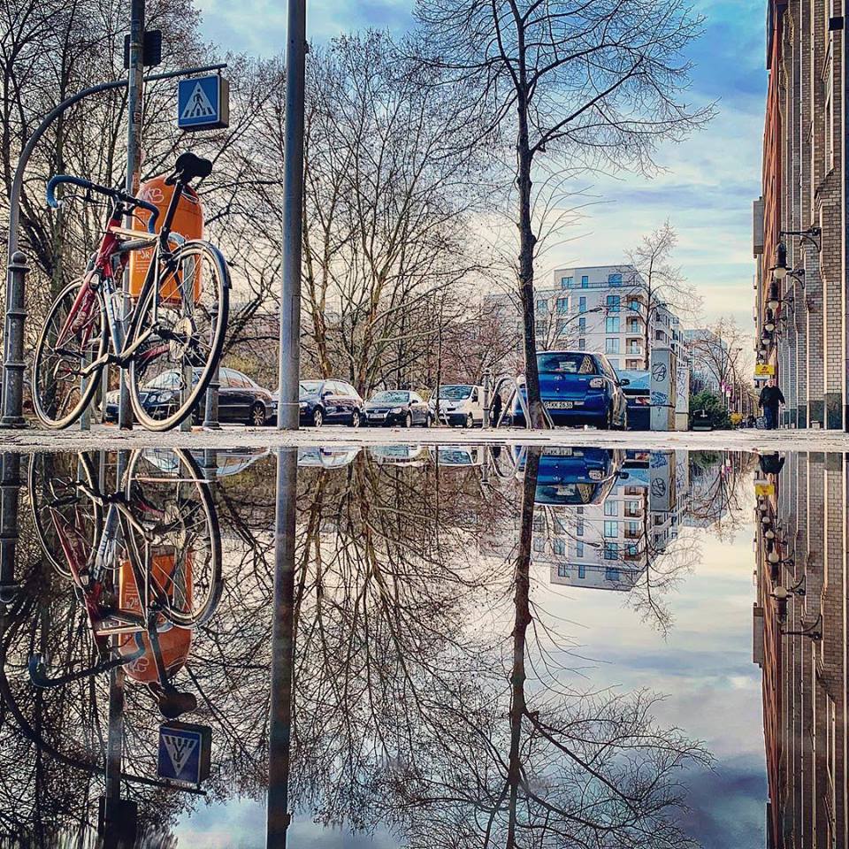 #paracegover Descrição para deficientes visuais: a imagem mostra o reflexo da calçada numa poça d’água: uma bicicleta parada ao lado de uma lixeira laranja; um carro azul, parte da fachada de um prédio e uma floresta de galhos secos. Depois de dias cinzentos, o céu acordou azul... — at Alnatura.